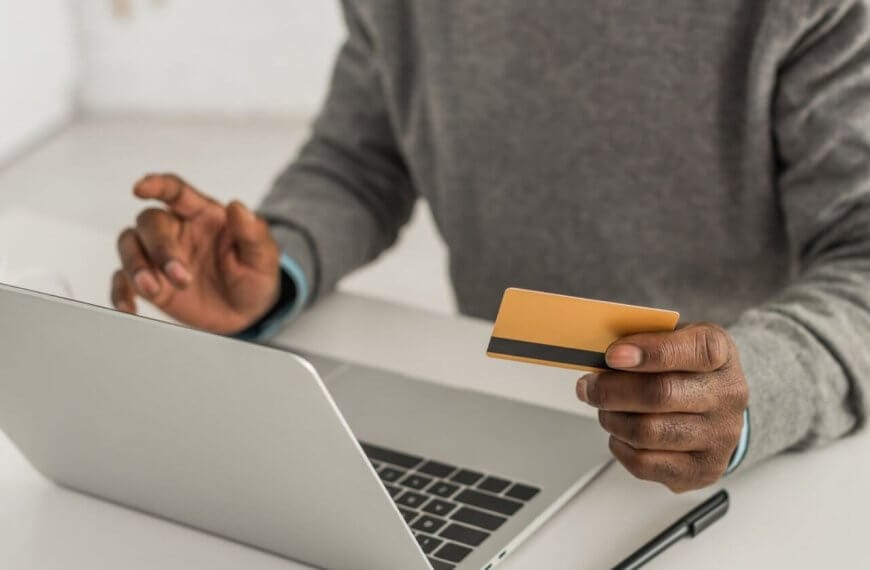 A person in a grey sweater holds a gold-colored credit card in their right hand while using their left hand to type on a laptop keyboard. They are likely engaged in online shopping, sitting at a white desk with a white background, and a pen is visible on the desk beside the laptop.