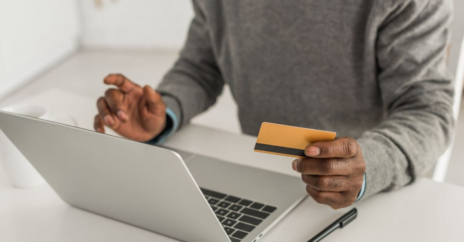 A person in a grey sweater holds a gold-colored credit card in their right hand while using their left hand to type on a laptop keyboard. They are likely engaged in online shopping, sitting at a white desk with a white background, and a pen is visible on the desk beside the laptop.