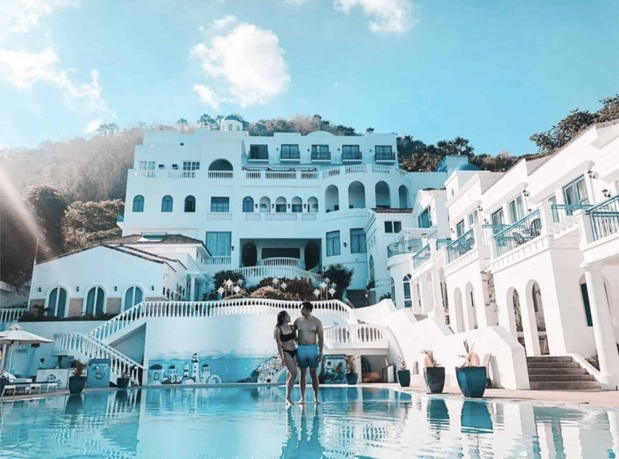 Couple stands by a large pool in front of a white Mediterranean-style resort with blue accents under a clear sky.