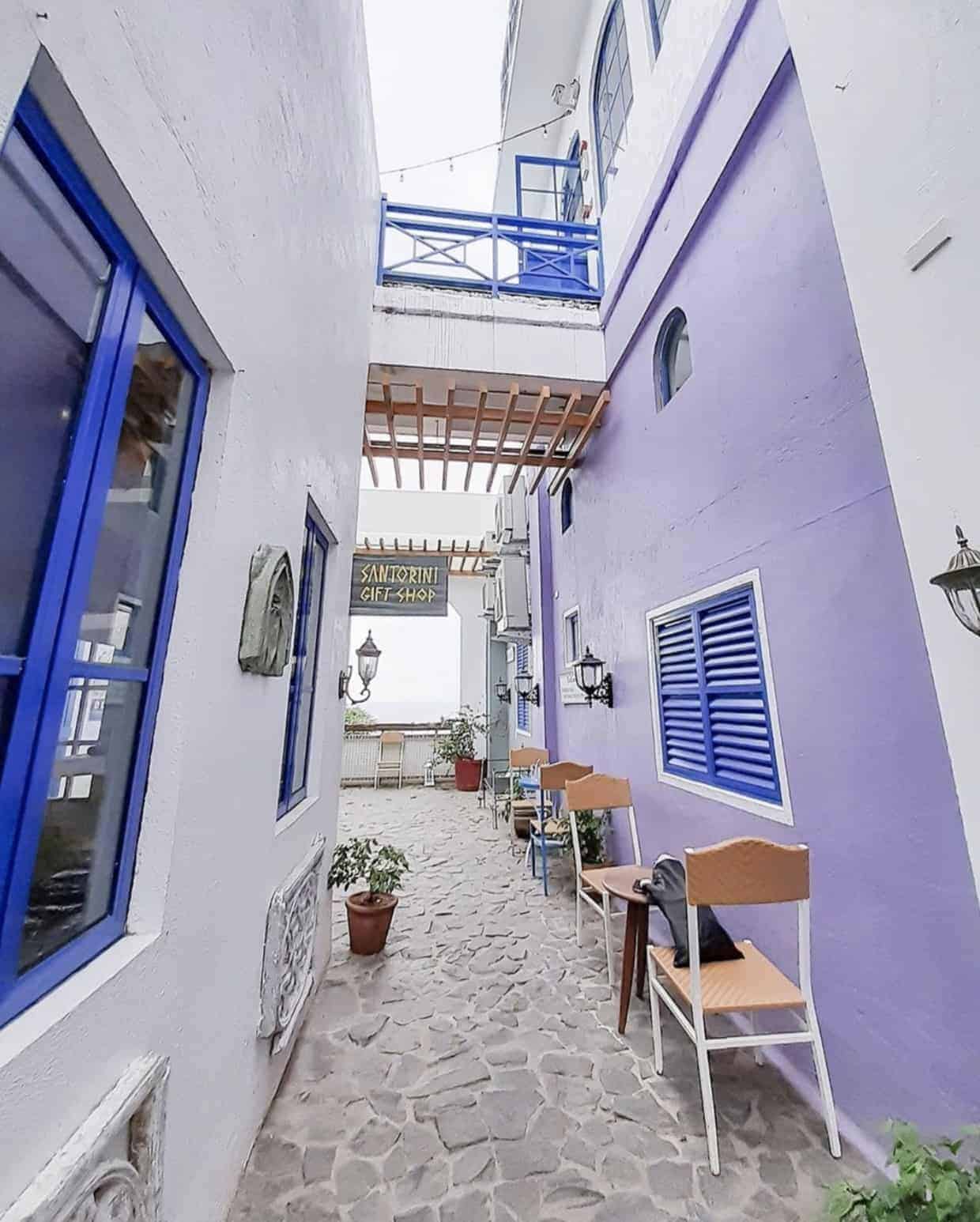 Narrow cobblestone alley with purple and white walls, blue windows, chairs, and a Santorini Gift Shop sign.