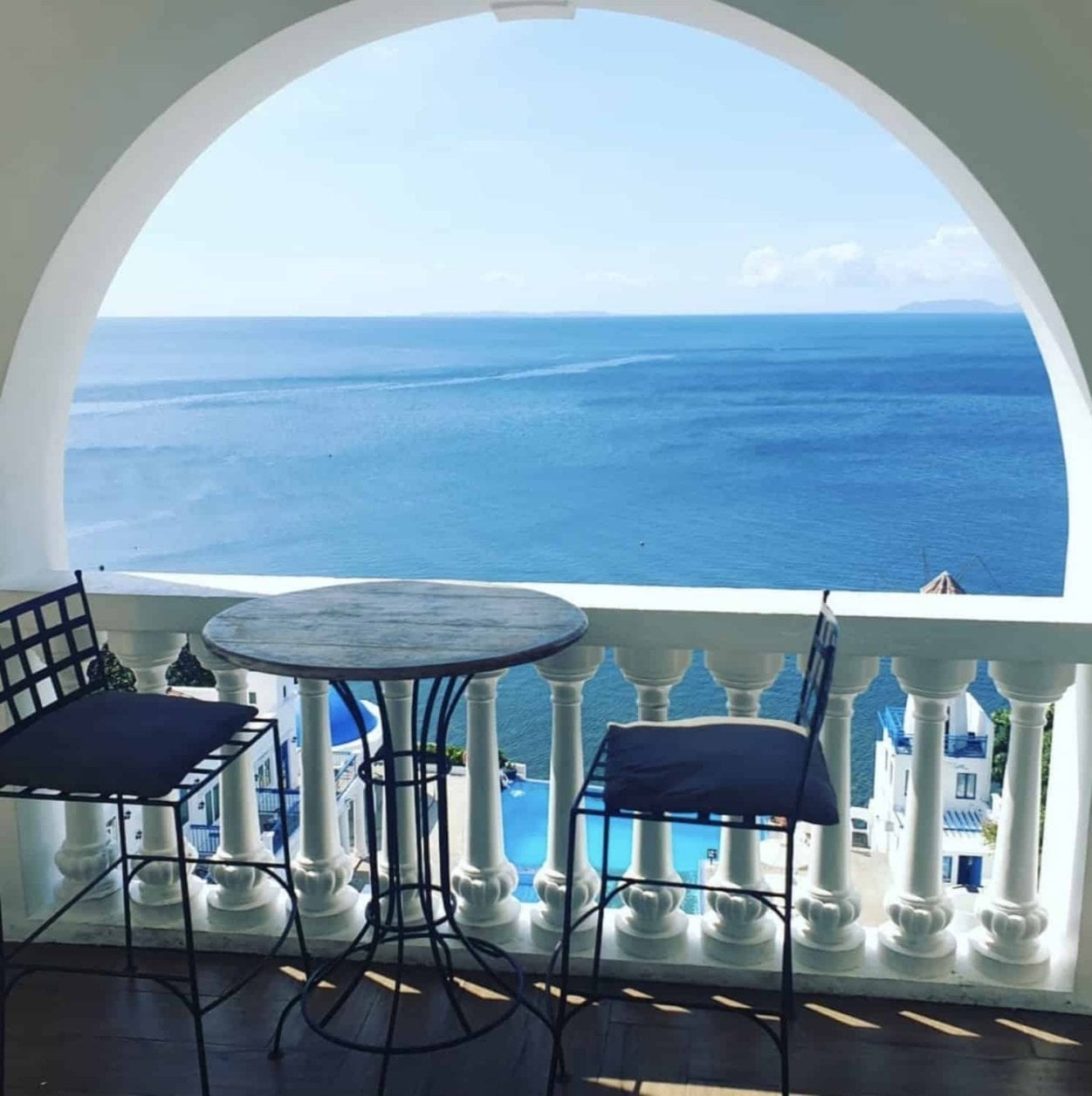 Balcony with a round table and two chairs overlooking a wide ocean view through a white archway.