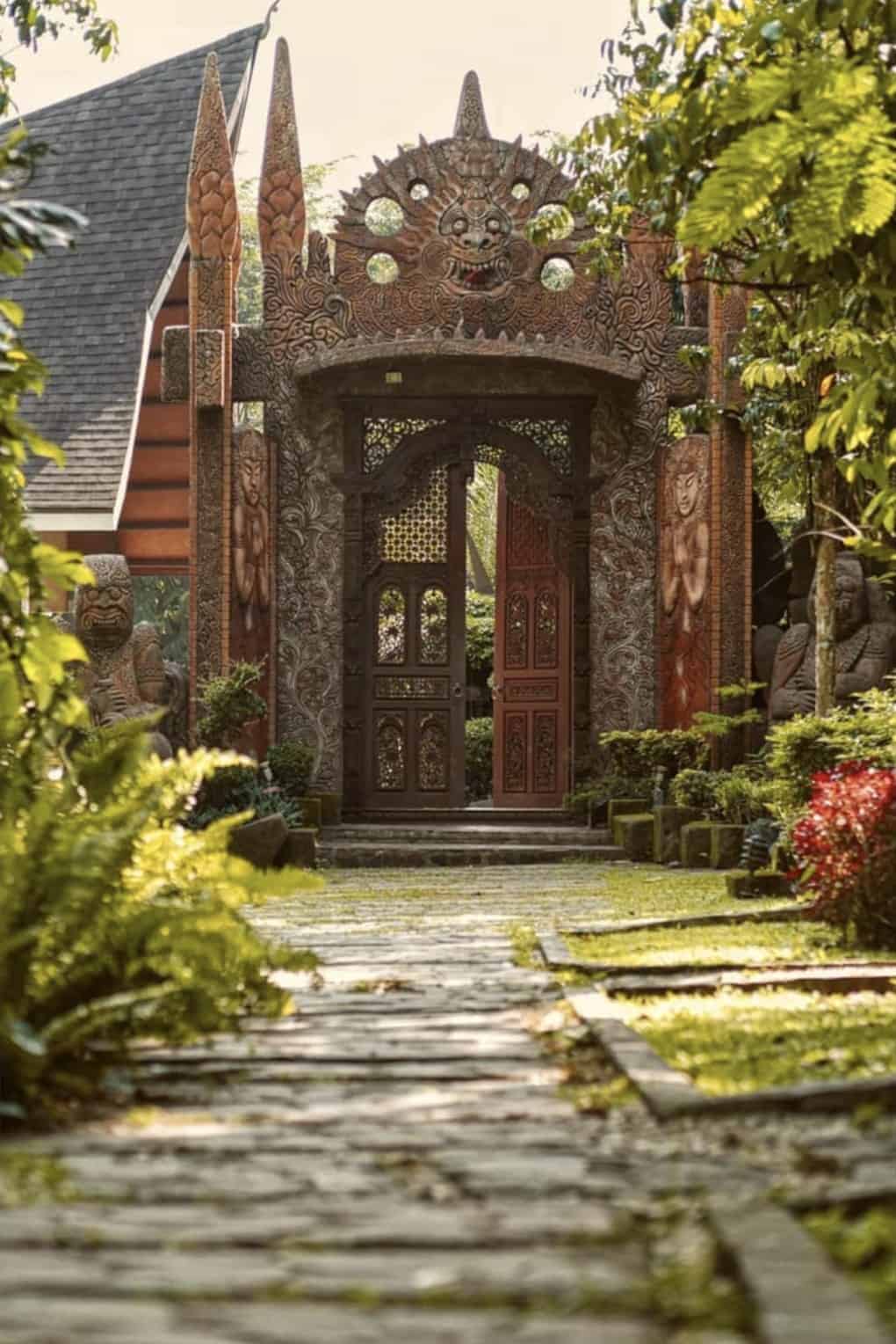 Ornate wooden gate with intricate carvings in a lush garden setting, featuring statues and vibrant green foliage.