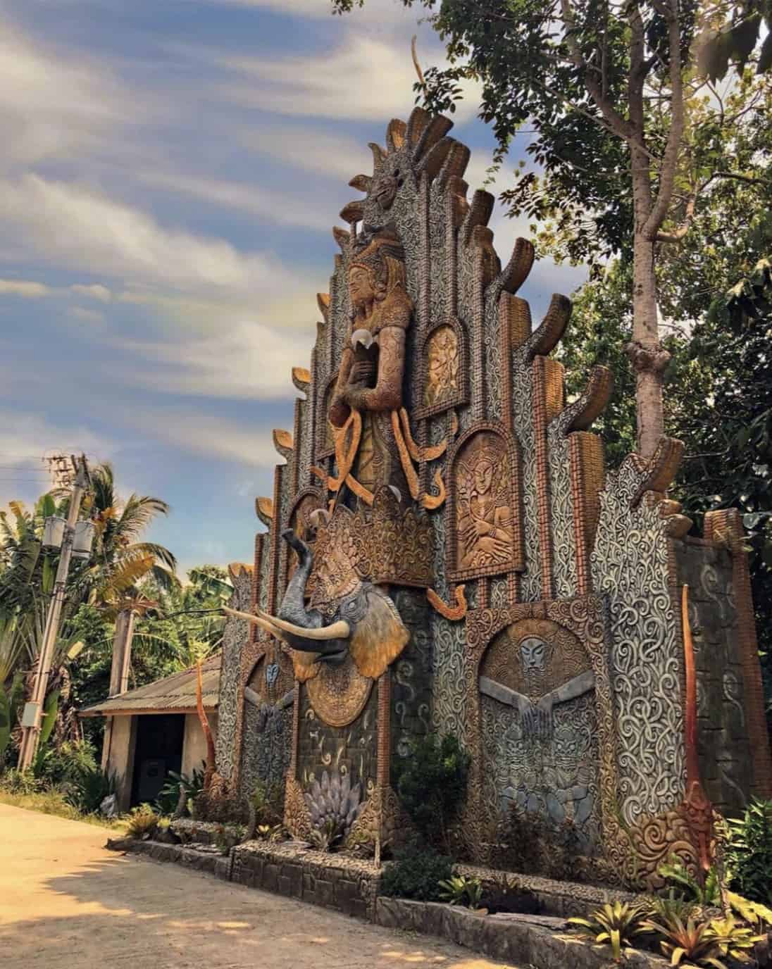 Ornate temple facade with intricate carvings, statues, and trees against a cloudy sky.