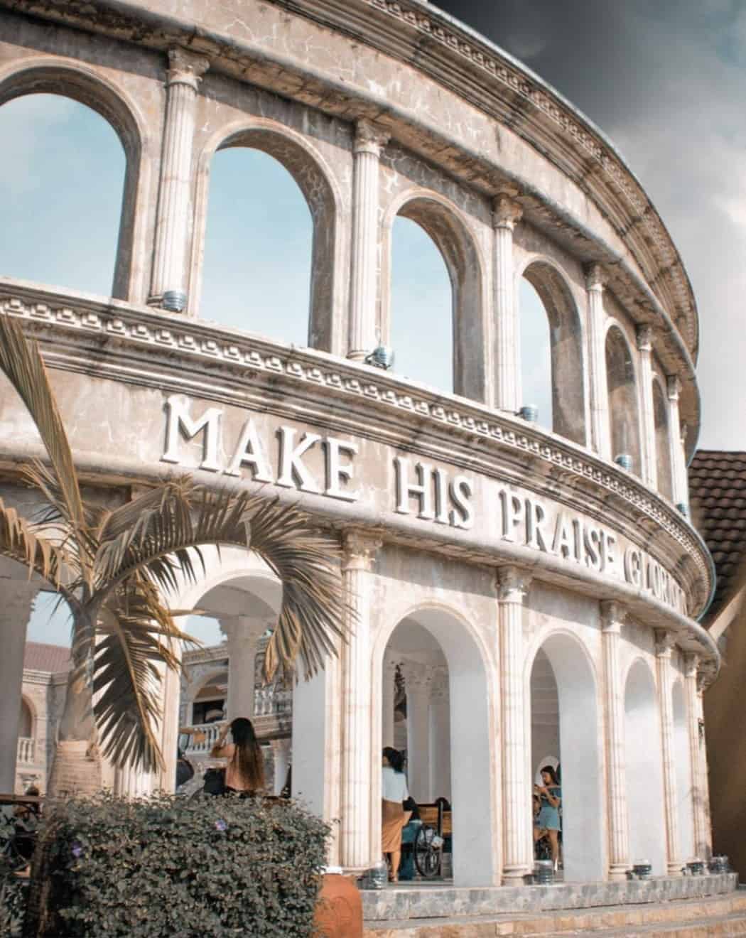 Colosseum-like structure with Make His Praise Glorious on the facade, under a cloudy sky.