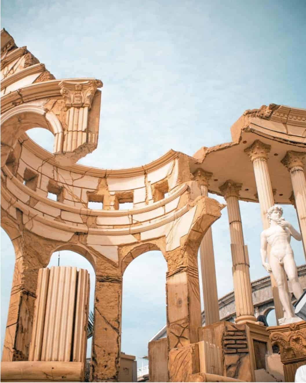 Ancient Roman-style ruins with columns, archways, and a statue under a blue sky.