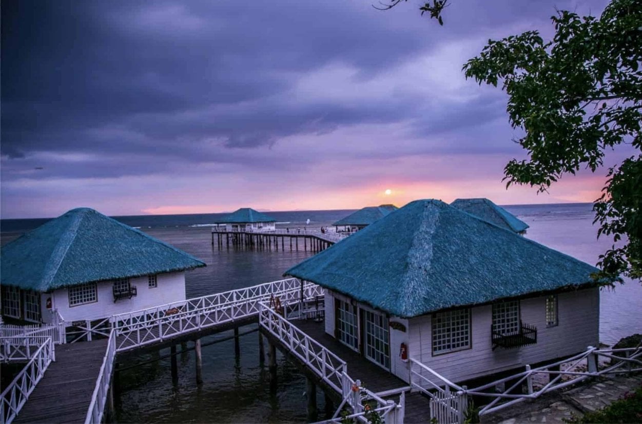 Overwater bungalows with blue roofs at sunset, under a dramatic cloudy sky, surrounded by calm sea waters.