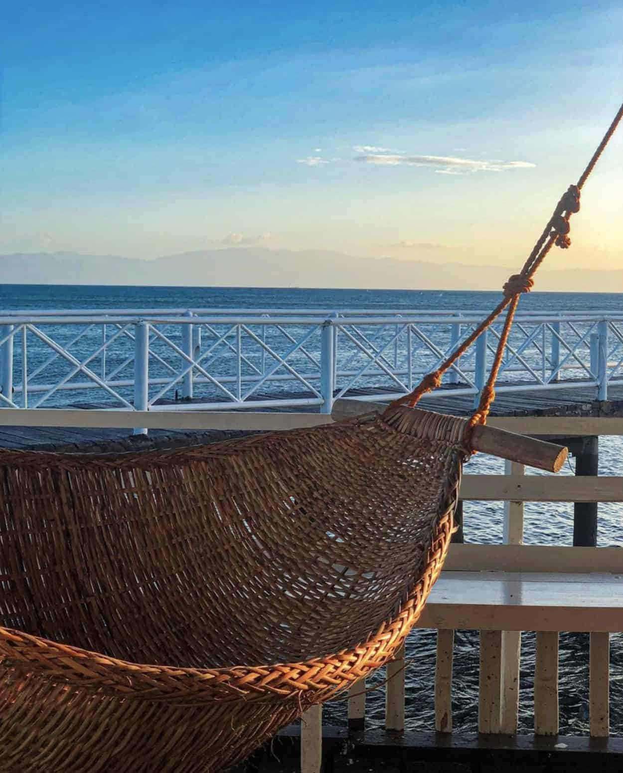 Woven hammock on a wooden deck by the sea at sunset, with a clear sky and mountains in the distance.