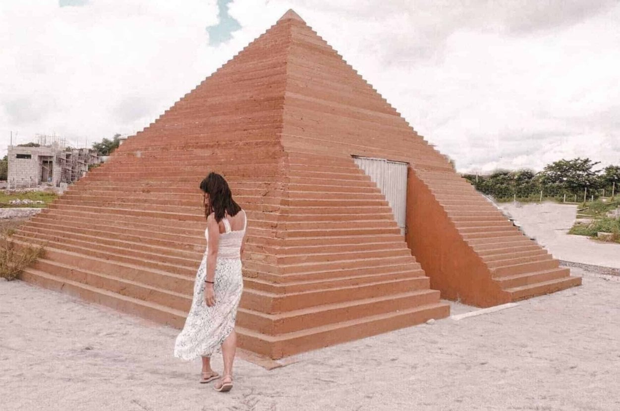 Woman in white dress walking by a small, red pyramid in a sandy outdoor setting under a cloudy sky.