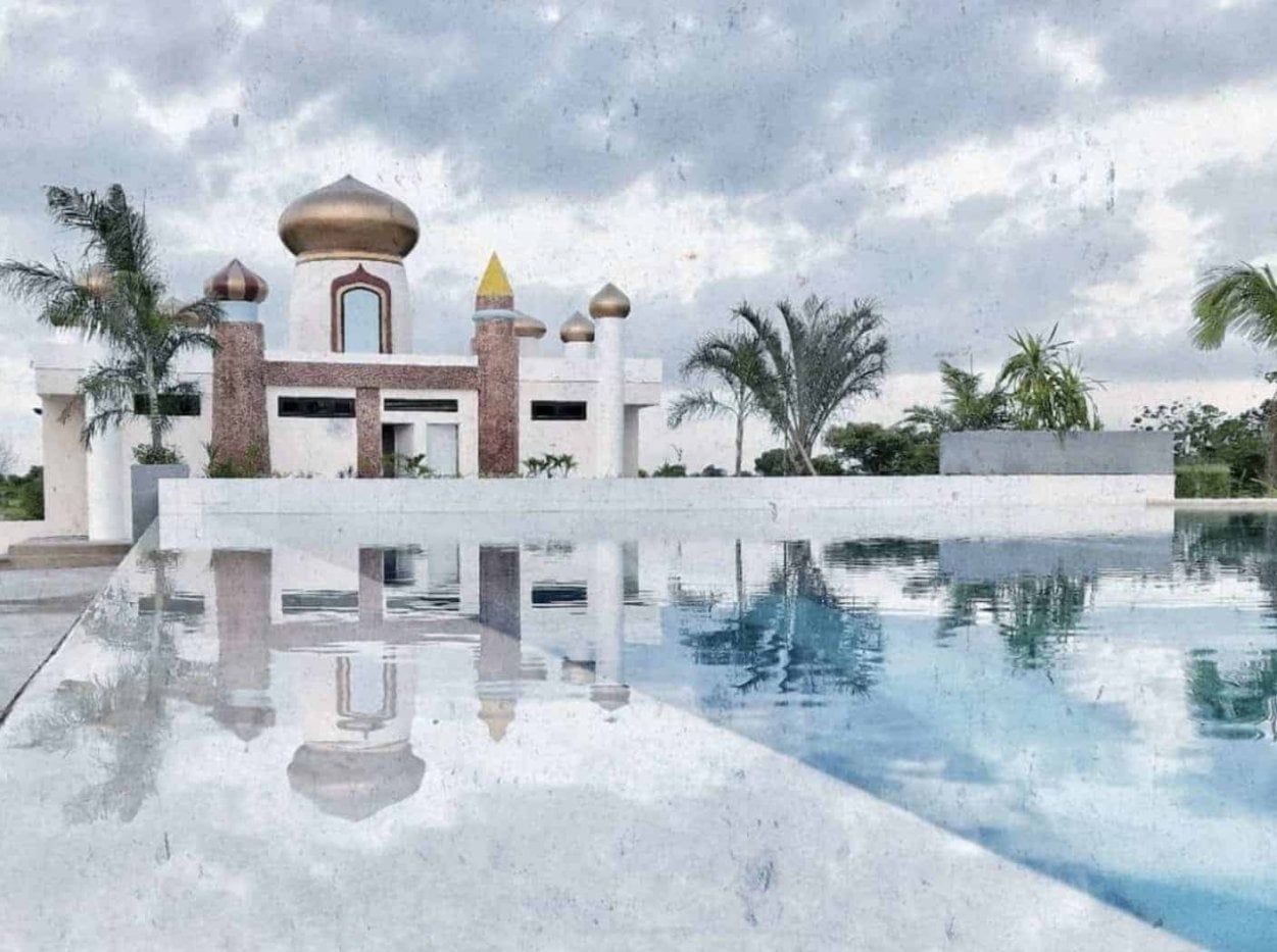 A serene pool reflects a building with golden domes, surrounded by palm trees under a cloudy sky.