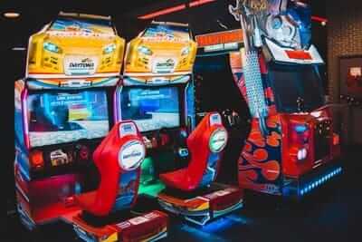 A vibrant arcade with Daytona USA racing games and a pinball machine, featuring colorful seats and neon lights.