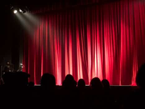 A dimly lit stage with closed red curtains and silhouetted audience in the foreground.