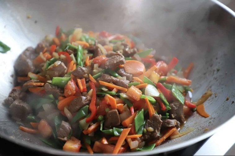 Sizzling stir-fry with beef, red peppers, carrots, green peas, and onions in a steaming wok.