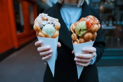 Person holding two bubble waffle cones filled with colorful ice cream and toppings.