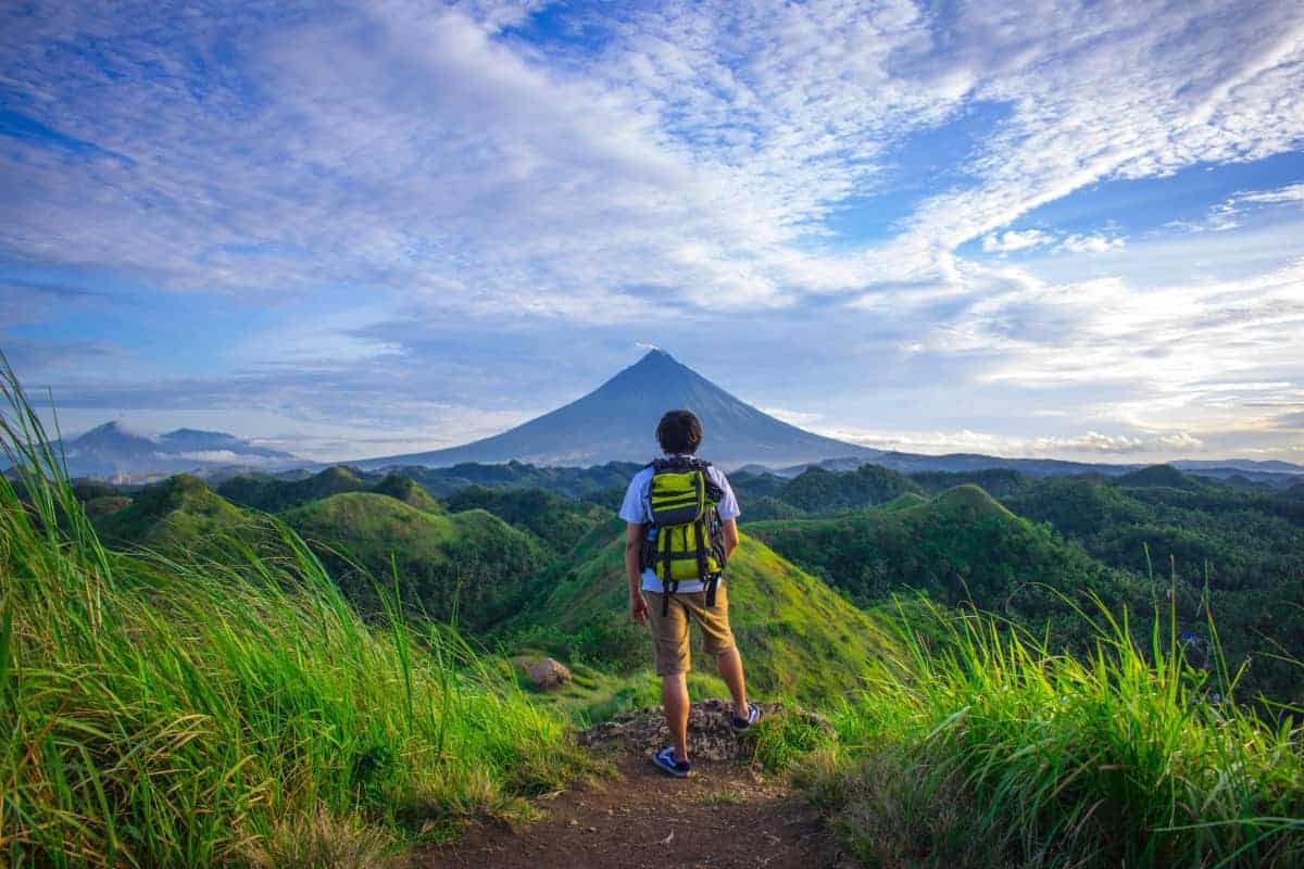 A person with a backpack stands on a grassy hill, facing a distant volcano beneath a blue sky with clouds.