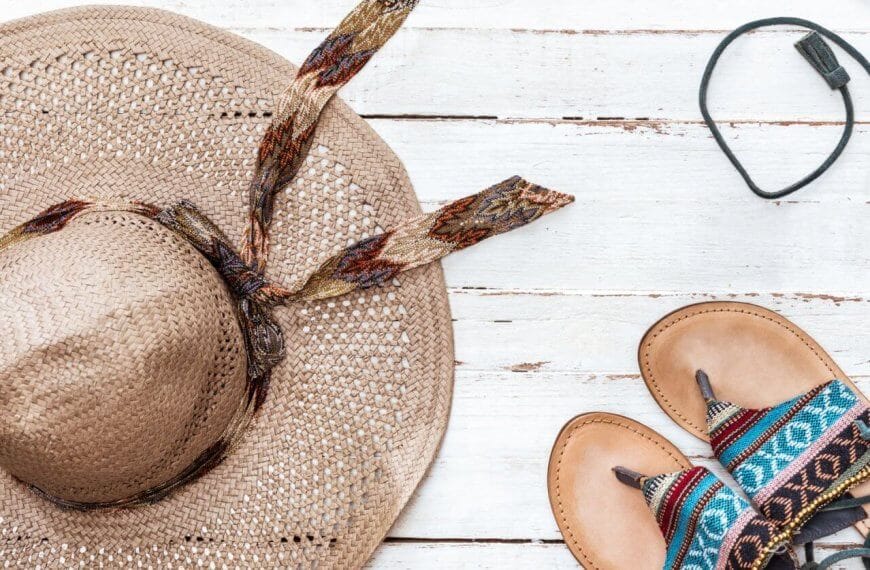 A straw hat with a patterned ribbon, a pair of leather sandals featuring colorful woven straps, and black earphones are artistically arranged on a white wooden surface. Evoking a casual, summery vibe perfect for Philippine holidays in 2024.