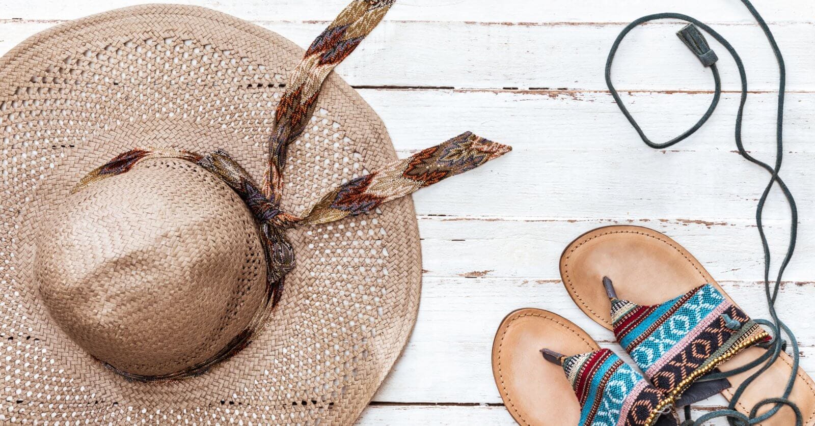 A straw hat with a patterned ribbon, a pair of leather sandals featuring colorful woven straps, and black earphones are artistically arranged on a white wooden surface. Evoking a casual, summery vibe perfect for Philippine holidays in 2024.
