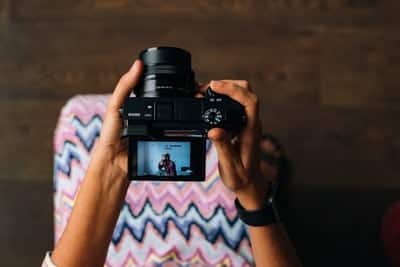 Hands holding a camera, displaying a patterned cloth and a person in the viewfinder, against a wooden floor.