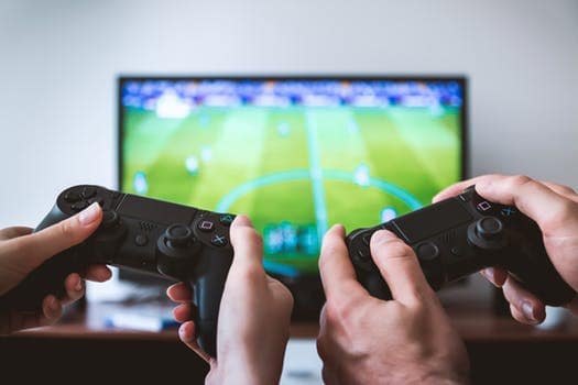 Two people holding black game controllers, playing a soccer video game on a TV screen in the background.