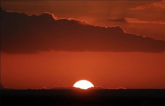 Sunset with a glowing orange sun partially hidden by dark clouds against a vibrant red sky.