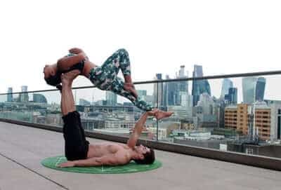 Two people practice acro yoga on a rooftop with a city skyline in the background.