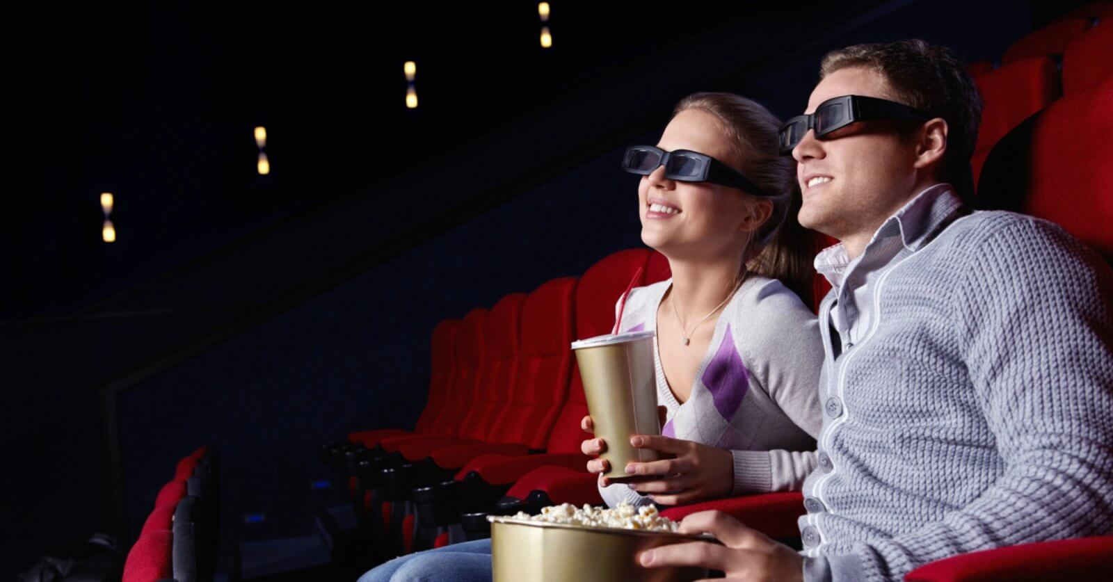 A couple wearing 3D glasses sits in a movie theater smiling and holding buckets of popcorn in red seats enjoying the magic of Pinoy movies come to life