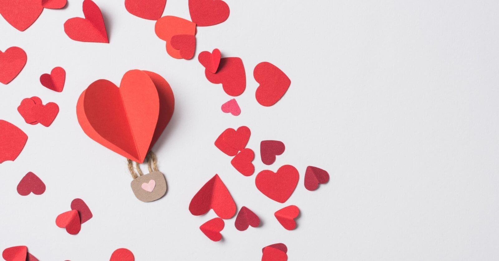 A paper hot air balloon with a red heart floats among scattered red and pink heart-shaped cutouts on a white background. Perfect for 2023 Valentine Gift Ideas, the whimsical scene features a brown basket with two strings, capturing love's playful essence.