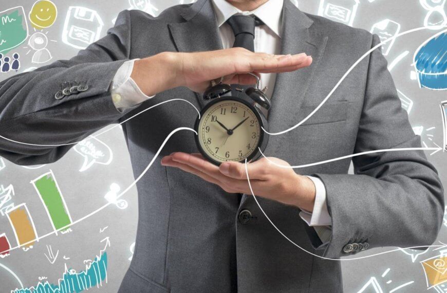 A man in a gray suit holds a traditional alarm clock with both hands against a background filled with various colorful doodles, including charts, graphs, a light bulb, a laptop, and other business-related symbols. The clock's hands point to 10:10. Lines connect the clock to the doodles, symbolizing 2023 business ideas.