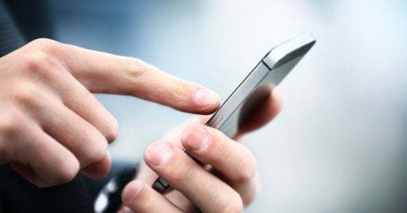 Close-up image of a person’s hands holding a sleek, new phone. The person is using their right index finger to touch the screen. The background is blurred with a mix of light and dark blue tones, focusing attention on the device and hands. The smartphone has a metallic edge, highlighting its modern design.