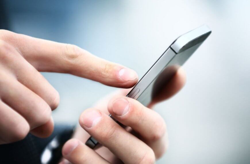 Close-up image of a person’s hands holding a sleek, new phone. The person is using their right index finger to touch the screen. The background is blurred with a mix of light and dark blue tones, focusing attention on the device and hands. The smartphone has a metallic edge, highlighting its modern design.
