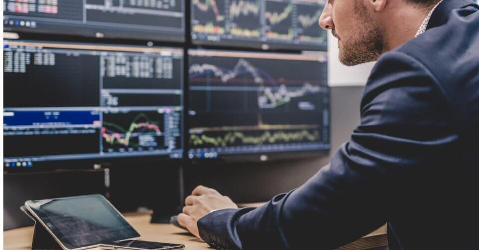a man in a suit sitting at a desk with several monitors, researching investment opportunities available through stock brokers in the philippines.