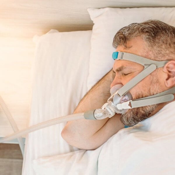 A man sleeps in a sunlit bedroom using a CPAP machine purchased from one of the top CPAP stores in Metro Manila. He lies on his side, with his head on a pillow, wearing a head strap connected to a nasal mask. A nightstand with a lamp is visible beside him.
