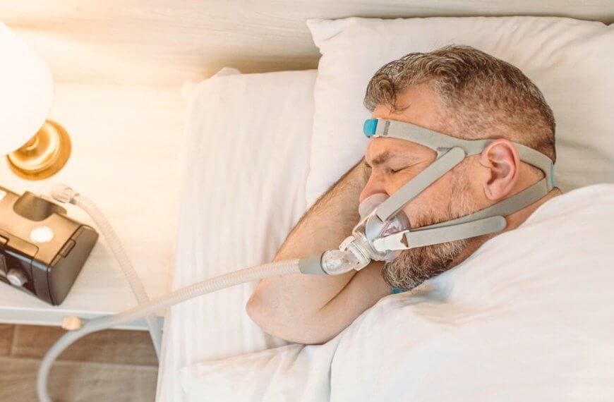 A man sleeps in a sunlit bedroom using a CPAP machine purchased from one of the top CPAP stores in Metro Manila. He lies on his side, with his head on a pillow, wearing a head strap connected to a nasal mask. A nightstand with a lamp is visible beside him.