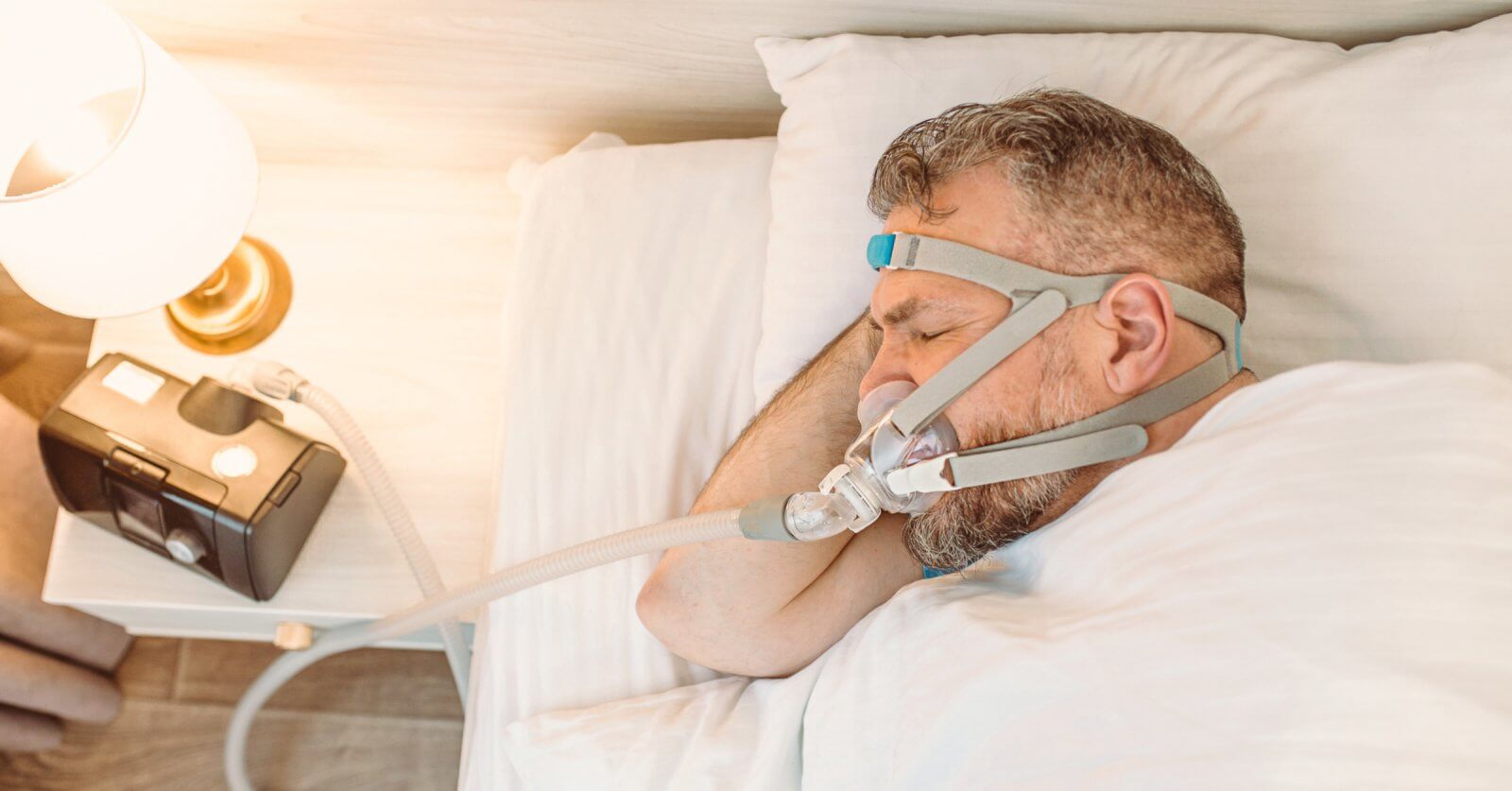 A man sleeps in a sunlit bedroom using a CPAP machine purchased from one of the top CPAP stores in Metro Manila. He lies on his side, with his head on a pillow, wearing a head strap connected to a nasal mask. A nightstand with a lamp is visible beside him.