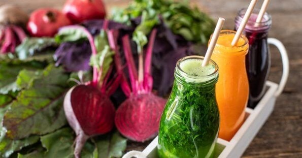 Three bottles of colorful vegetable juices—green, beetroot red, and orange—from top juice bars in Metro Manila, placed on a wooden table, surrounded by fresh vegetables like beets, carrots, and leafy greens.
