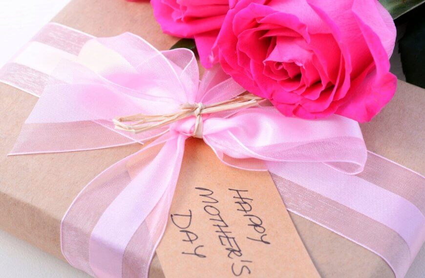 A close-up of a gift wrapped in brown paper tied with a pink ribbon. A tag reading "Happy Mother's Day" is attached to the ribbon, and two bright pink roses rest beside the gift, perfect for celebrating Mother's Day.
