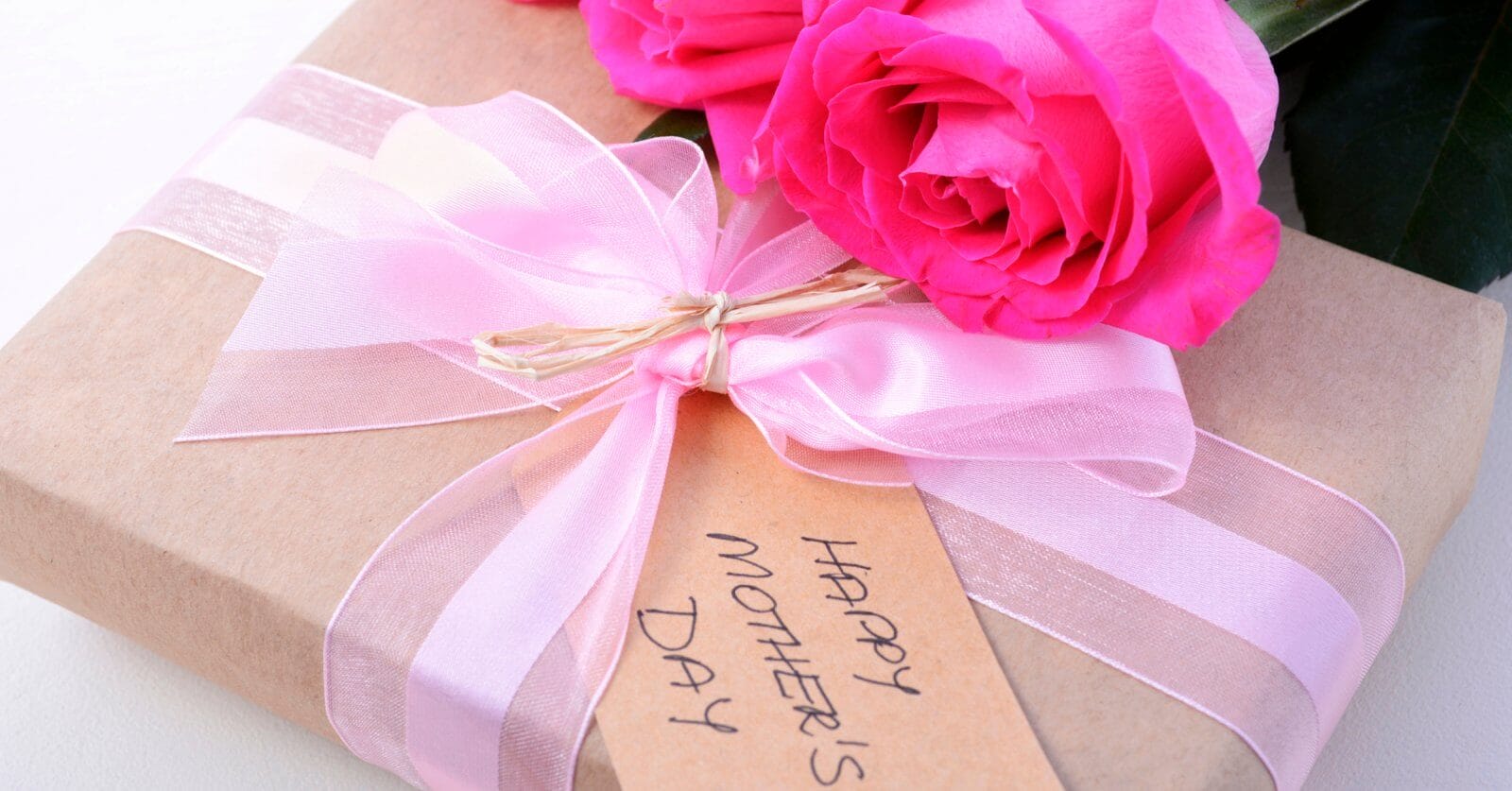 A close-up of a gift wrapped in brown paper tied with a pink ribbon. A tag reading "Happy Mother's Day" is attached to the ribbon, and two bright pink roses rest beside the gift, perfect for celebrating Mother's Day.