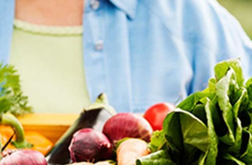 A person holding a wooden basket full of vibrant fresh vegetables including red apples, green lettuce, and purple beets, all sourced from organic hotspots, with a focus on healthy eating. The person wears a light green shirt and blue jacket.