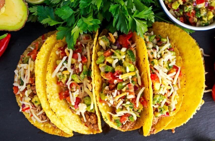 Three crunchy tacos filled with seasoned ground beef, diced tomatoes, chopped onions, shredded cheese, and garnished with cilantro. The tacos are placed on a dark surface surrounded by halved avocados, red chili peppers, a bowl of salsa, and a bunch of cherry tomatoes. Discover the best taco places inspired by Taco Places Metro Manila.