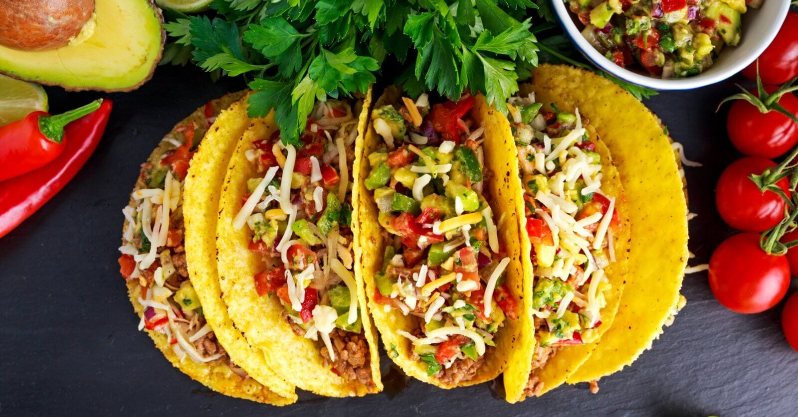 Three crunchy tacos filled with seasoned ground beef, diced tomatoes, chopped onions, shredded cheese, and garnished with cilantro. The tacos are placed on a dark surface surrounded by halved avocados, red chili peppers, a bowl of salsa, and a bunch of cherry tomatoes. Discover the best taco places inspired by Taco Places Metro Manila.