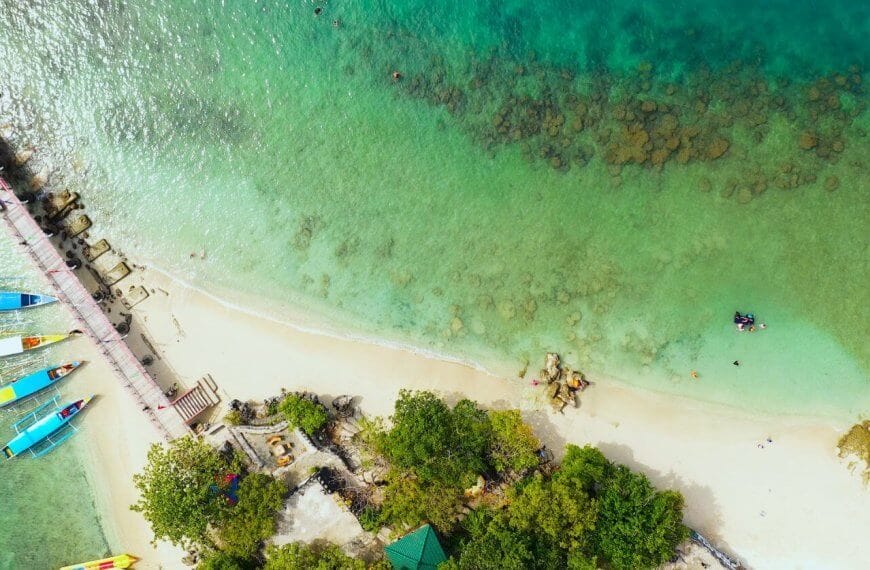 Aerial view of Alaminos City beach with turquoise waters and a thin line of white sand. A pier extends from the shore on the left, near colorful boats docked. Swimmers are visible in the clear water, with patches of rocks observable underwater. Lush green trees line the edge, perfect for post-swim visits to must-try restaurants nearby.
