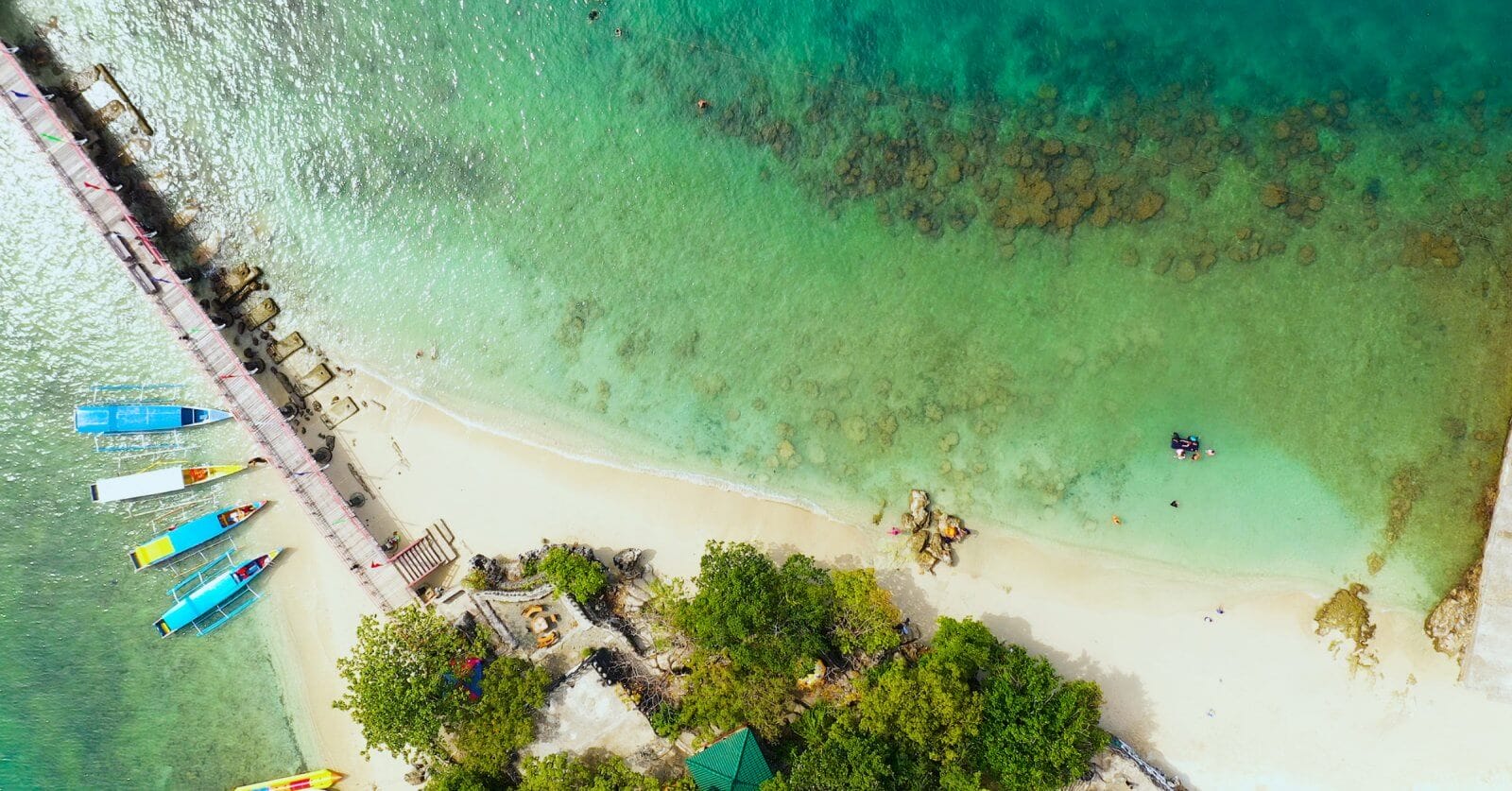 Aerial view of Alaminos City beach with turquoise waters and a thin line of white sand. A pier extends from the shore on the left, near colorful boats docked. Swimmers are visible in the clear water, with patches of rocks observable underwater. Lush green trees line the edge, perfect for post-swim visits to must-try restaurants nearby.