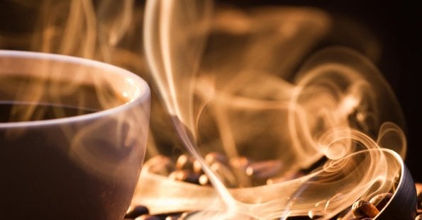 A white cup filled with steaming hot coffee sits on a surface scattered with coffee beans at a Manila 24/7 coffee shop. The steam rises in delicate, swirling patterns, creating an artistic effect in the warm, ambient lighting. The focus is close-up, capturing the texture of the beans and the ethereal quality of the steam.