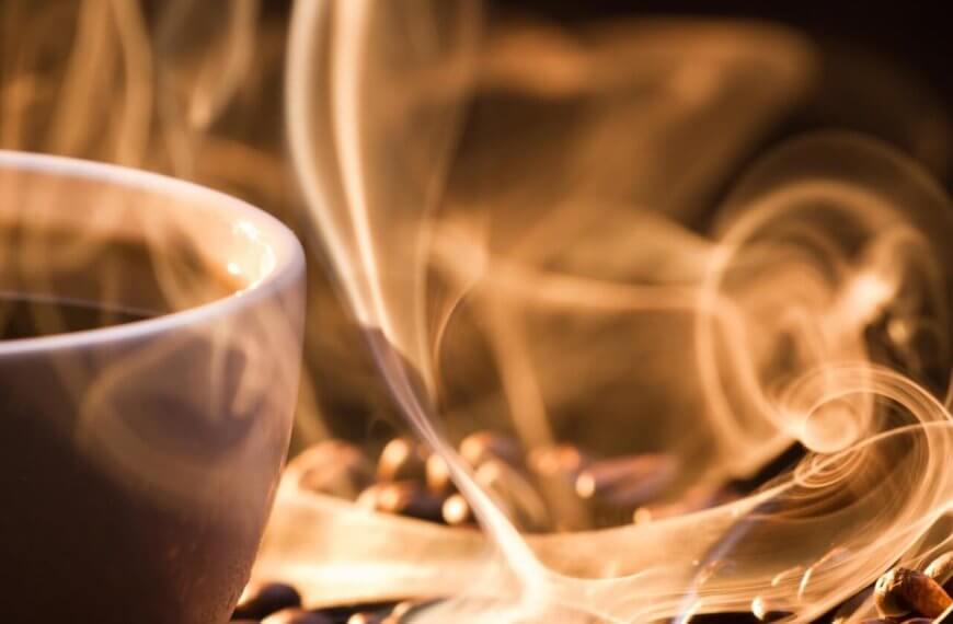 A white cup filled with steaming hot coffee sits on a surface scattered with coffee beans at a Manila 24/7 coffee shop. The steam rises in delicate, swirling patterns, creating an artistic effect in the warm, ambient lighting. The focus is close-up, capturing the texture of the beans and the ethereal quality of the steam.