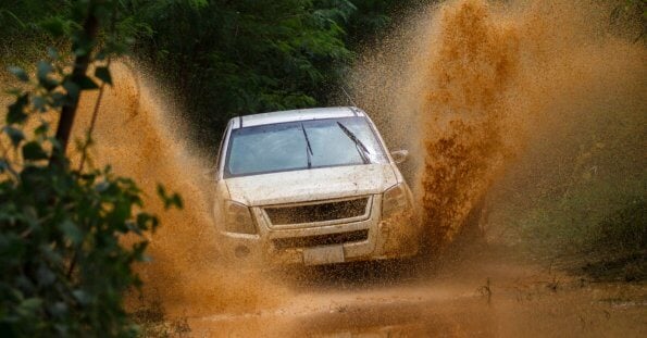 A white SUV drives through a muddy terrain, splashing mud high into the air. The vehicle is surrounded by lush green foliage, showcasing the adventure and rugged environment. The dynamic shot captures the energy of visiting Metro Manila's best 4x4 offroad shops and experiencing an exhilarating off-road journey.