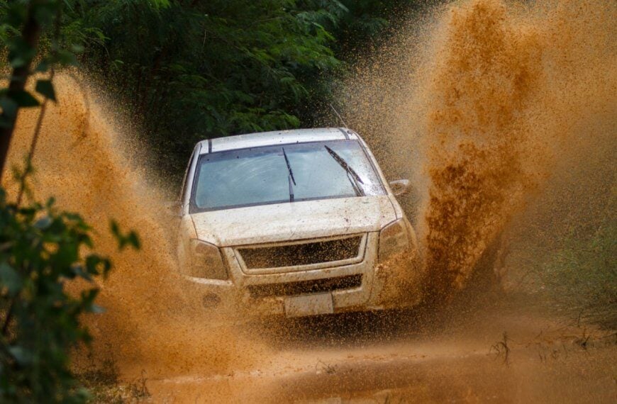 A white SUV drives through a muddy terrain, splashing mud high into the air. The vehicle is surrounded by lush green foliage, showcasing the adventure and rugged environment. The dynamic shot captures the energy of visiting Metro Manila's best 4x4 offroad shops and experiencing an exhilarating off-road journey.