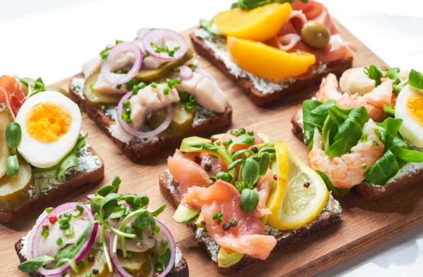 A wooden board topped with six open-faced sandwiches, a must-try at Sulit Restaurants in El Nido, featuring colorful toppings like sliced boiled eggs, pickles, red onions, various greens, salmon, shrimp, lemon slices, peach slices, and olives on dark bread. The sandwiches are neatly arranged in two rows of three.