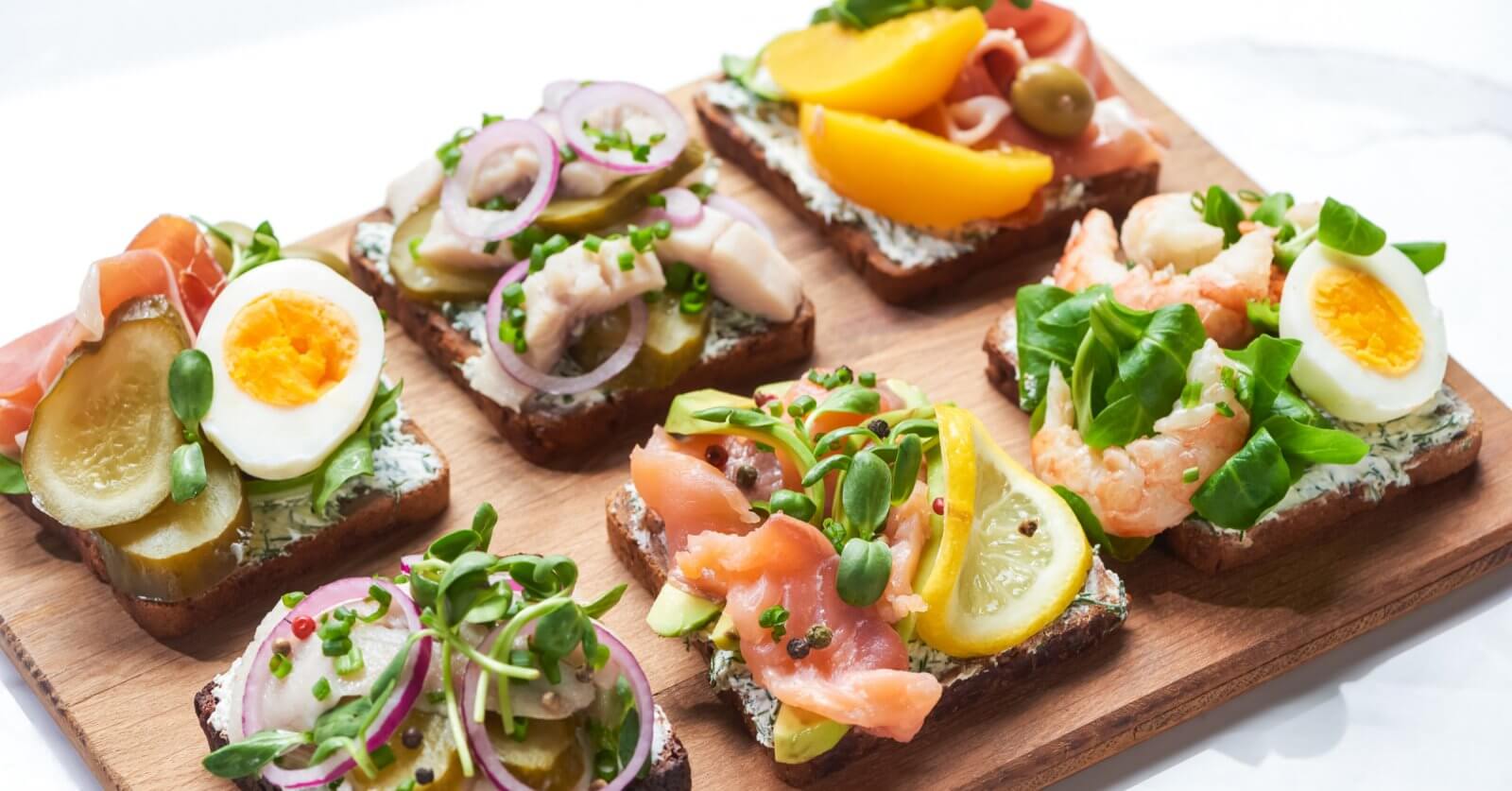 A wooden board topped with six open-faced sandwiches, a must-try at Sulit Restaurants in El Nido, featuring colorful toppings like sliced boiled eggs, pickles, red onions, various greens, salmon, shrimp, lemon slices, peach slices, and olives on dark bread. The sandwiches are neatly arranged in two rows of three.
