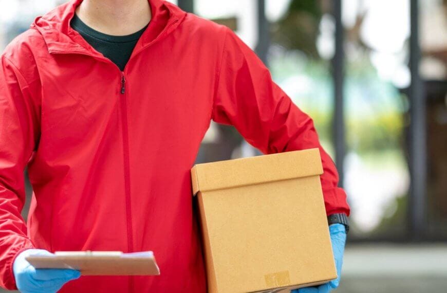A person wearing a red jacket and blue gloves holds a brown cardboard box in one hand and a clipboard in the other. The background is slightly blurred, showing an indoor setting with large glass windows and greenery outside, possibly indicating J&T Drop-Off Locations within Metro Manila, emphasizing convenience.