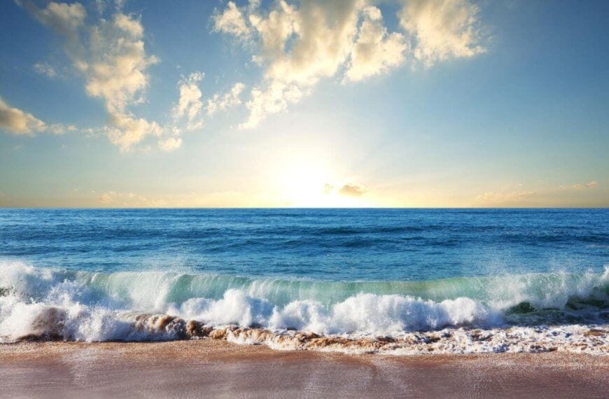 A scenic view of a Zambales beach at sunrise with gentle waves lapping the shore. The golden sunlight illuminates the sky, casting a warm glow on scattered clouds. The calm ocean stretches to the horizon, blending into the sky where the sun peeks just above the water's edge—truly one of the best Zambales resorts.