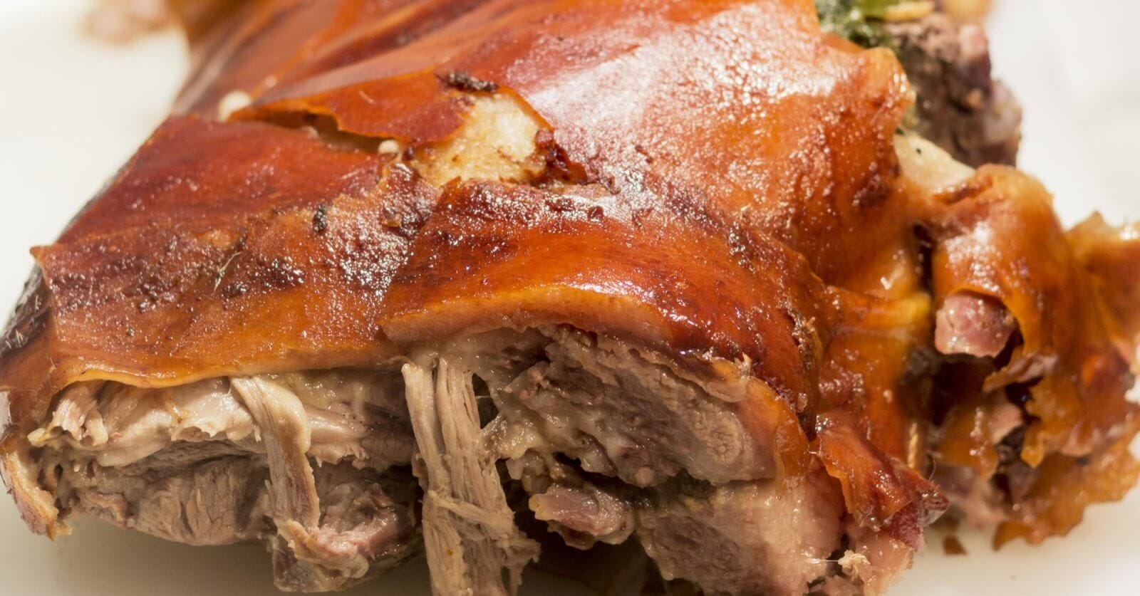 A close-up of a roasted pork dish from one of the best restaurants in Alabang, showcasing crispy, golden-brown skin with some tender, juicy shredded meat visible underneath. The meat appears well-cooked and succulent, with a slightly charred, flavorful exterior. The plain white background highlights the dish's texture.
